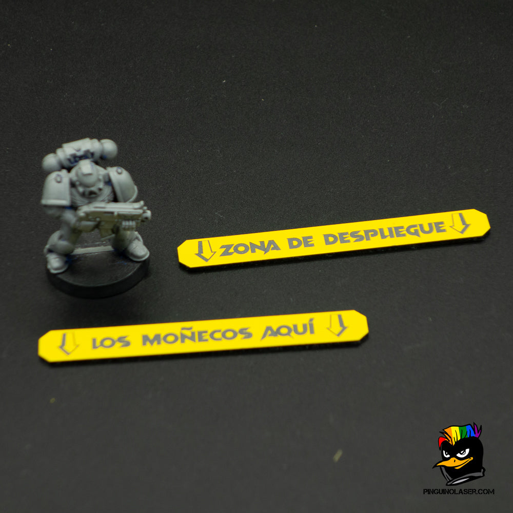 Foto de marcadores de despliegue para miniaturas con una miniatura de referencia. Los marcadores son rectangulares, amarillos con grabado en negro. Uno reza "zona de despliegue" y otro "los moñecos aquí".