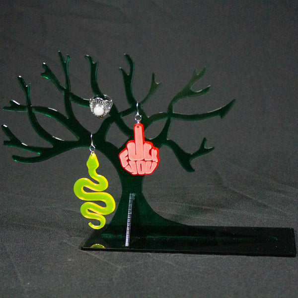 Joyero en forma de árbol sin hojas en color verde oscuro. Cuelgan pendientes y un anillo de las ramas. Un pendiente tiene forma de serpiente y el otro tiene las palabras "fuck you".